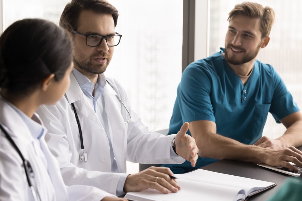 Ein selbstbewusster junger Chefarzt leitet eine Besprechung am runden Tisch mit einem vielfältigen Team von medizinischen Fachkräften, um Patientenfälle zu diskutieren.